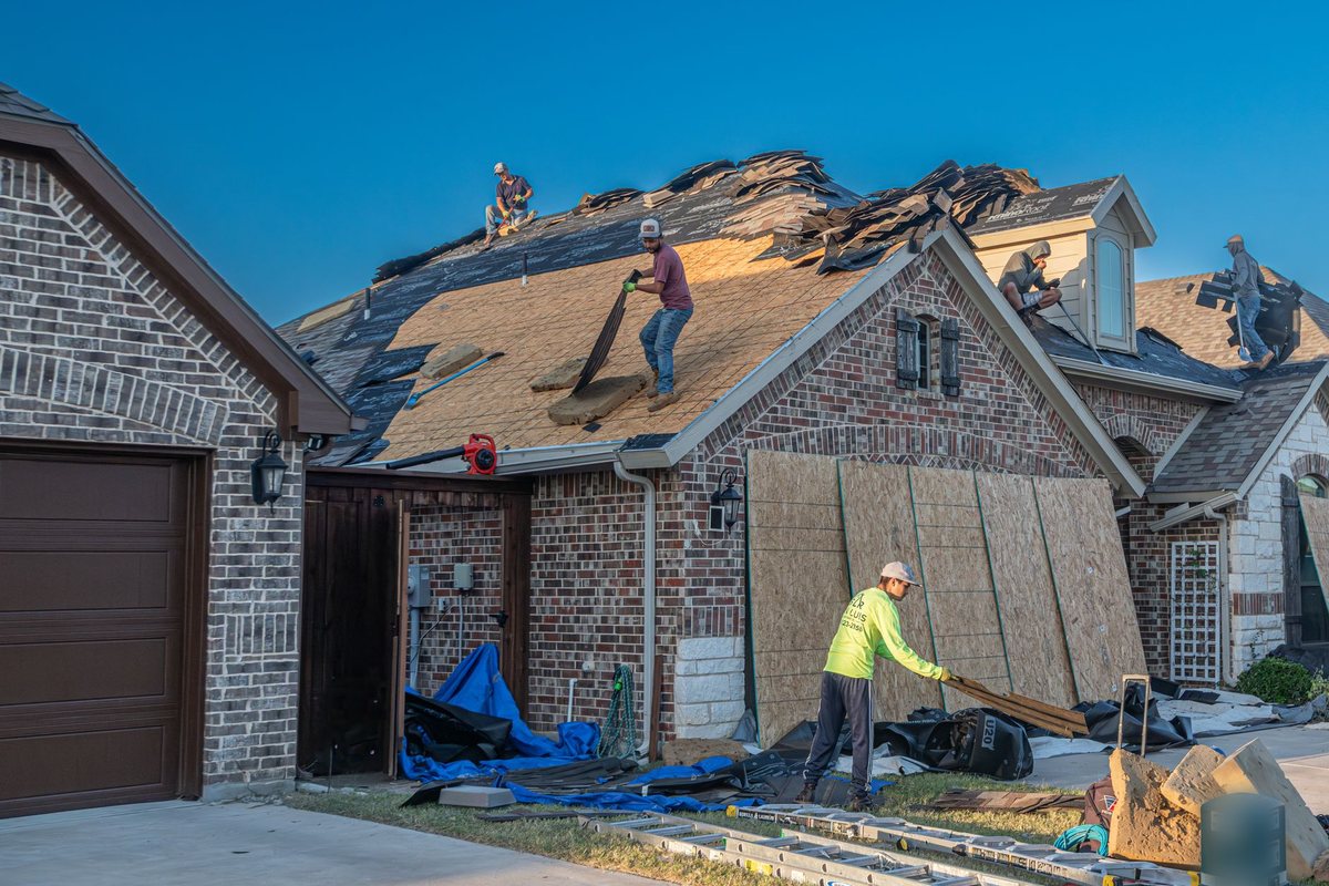 A crew tearing off old shingles from a brick house, plywood stacked in the driveway